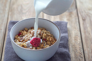 A plate of muesli stands on a wooden table. Someone pours cream into muesli. selective focus