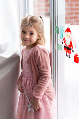Vertical view of beautiful smiling little girl with blond hair and brown eyes in dressy clothes and tiara holding a a star wand next to a window © Anne Richard