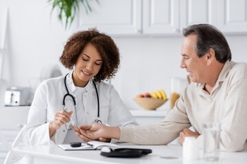 smiling african american doctor taking blood sample of mature patient with lancet pen