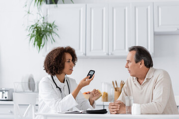 side view of curly african american doctor holding glucometer device and talking with middle aged patient
