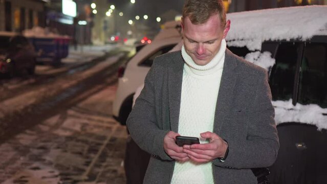 Man send sms text messages using app on smartphone outdoors under snowfall near passing by car at night. Posting social media, online shopping, texting, e commerce