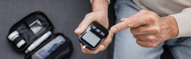 partial view of middle aged man with diabetes pointing at glucose meter device and sitting on sofa, banner