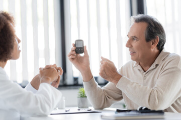 Positive mature patient holding glucometer near blurred african american doctor in clinic