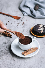Pouring cocoa powder into hot drink, served with muffin and cinnamon isolated on gray background. 