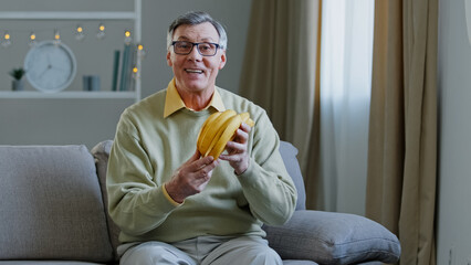Caucasian old man aged 60s angry man sits on sofa at home talking looking at camera blog male nutritionist gastroenterologist holds yellow fruits ripe sweet bananas advises healthy food