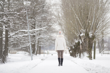 Young adult woman slowly walking on white snow covered sidewalk at park in cold winter day. Spending time alone. Enjoying peaceful stroll. Back view.