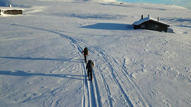A couple is skiing from their cabin at the mountain. It is winter in norway and it is a lovely day with a clear blue sky. They are cross-country skiing down the mountain.