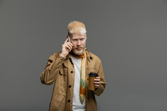 bearded albino man in shirt jacket holding paper cup and talking on smartphone isolated on grey