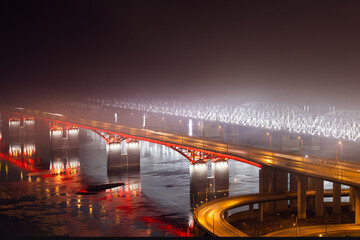 Fototapeta na wymiar Night cityscape with two illuminated bridges across the Yenisei river disappearing into thick fog in Krasnoyarsk, Russia. Industrial smog in a city with bad ecology.Backlight road and railway bridges