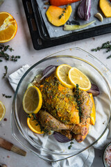 Raw spring chicken (coquelet jaune) in French herbs, orange zest and butter marinade in glass baking dish and raw vegetables on baking tray.