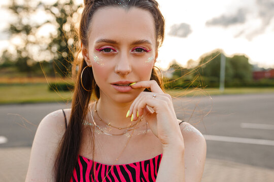 Portrait of confident young female teenager with glitter on face looking at camera in street at sundown 