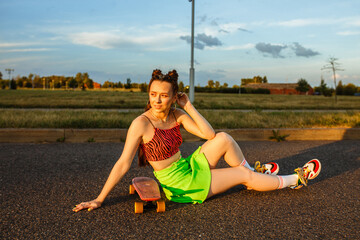 Full length of young female hipster sitting with skateboard on road at sunset in summer and looking...