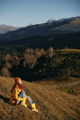 Fototapeta premium Woman full-length sitting resting on a hill smiling with teeth and looking at the mountains in a yellow raincoat and jeans happy camping trip in the spring, freedom lifestyle 