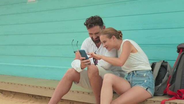 Young restful couple sitting on wooden bench by azure house wall, looking through their new photos in mobile phone taken on the beach and discussing one of them