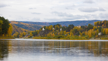 Autumn panoramic landscape on the Mana river with yellow trees and mountains, Russia. Pavilions and houses on the steep bank of the river. Cloudy day
