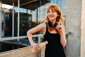 Ginger woman with headphones smiling and drinking water after workout