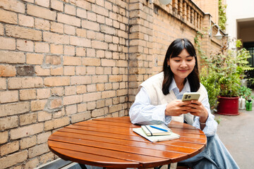 Fototapeta na wymiar Smiling young asian girl using smartphone while sitting in cafe outdoors