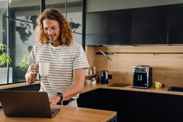 Young man drinking coffee while working on laptop in office kitchen