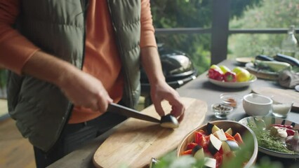 Cropped shot of hands of male chef cutting eggplant and putting pieces into bowl with veggies while cooking food on outdoor terrace