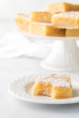 A lemon square with a bite out, with more on a dessert stand in behind.