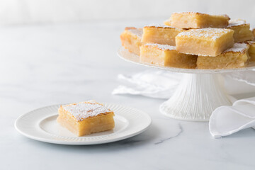 A lemon bar on a plate with a pedestal stand full of them in behind.