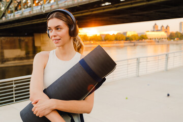 Young sportive woman holding yoga mat while standing on embankment