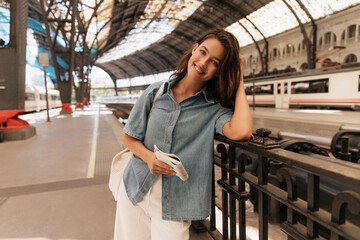 Positive young caucasian girl posing looking at camera spends time at railway station alone. Brunette wears casual clothes and bag. Concept of tourism.