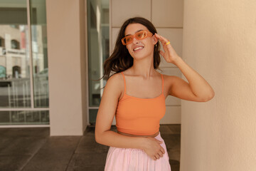 Beautiful young caucasian woman looks away smiling corrects her hair outside. Brunette wears top, pants and sunglasses. Concept of leisure vacation.