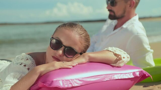Young serene woman in sunglasses keeping head on pink inflatable mattress while sunbathing on tropical beach on hot sunny day against her husband relaxing on seashore