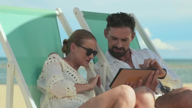 Young man with notepad commenting photos during slide show while sitting on deckchair next to his wife in sunglasses lisrtening to him and looking at tablet screen