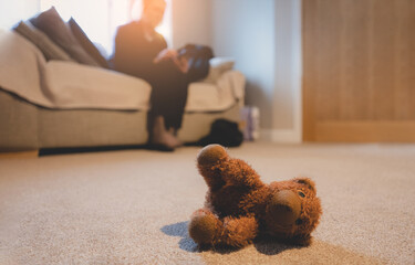 Teddy bear lying on carpet with blurry woman sitting on sofa,Lonely teddy bear lay down alone in living room, lonely concept, Domestic Family violence, International missing children's day.