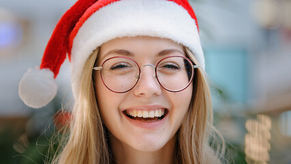 Close up portrait funny smiling toothy laughing happy Caucasian woman girl lady wearing Santa Claus Christmas hat and eyeglass looking at camera smile laugh headshot female face New Year celebration