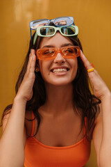 Positive young caucasian girl in three pair of sunglasses looks away with orange wall in background. Brunette wears orange top. Concept of positive moments.