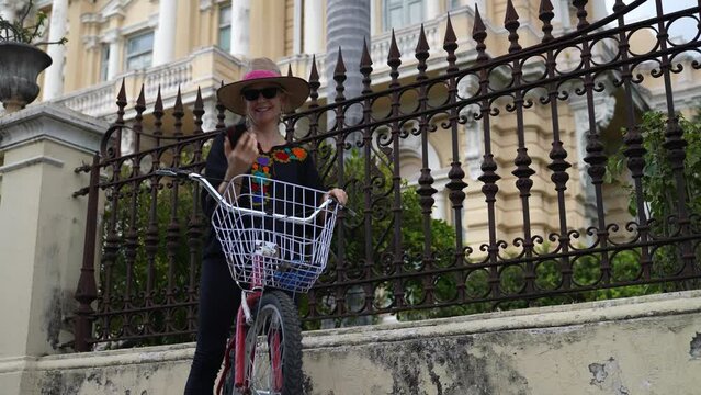 Pretty, mature elderly woman in ethnic clothing on bike taking selfie with smart phone on the Montejo Central Avenue with museums, restaurants, mansions in Merida, Yucatan, Mexico.