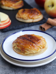 A plate of classic traditional mini French apple tart thin (tarte fine aux pommes) with its sliced fruit, cinnamon in background. It made with butter puff pastry pie, caramelized with brown sugar.