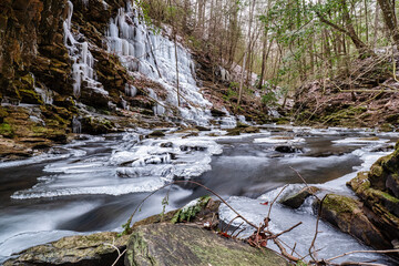 An icy mountain stream by a hiking trail with moss covered rocks and icicles hanging from bluff ....
