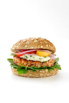 Salmon burger on a whole wheat bun with lettuce, red onion, red tomatoes, yellow tomatoes, sauce, and fresh dill isolated on a white background.