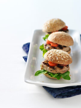Three grilled turkey burger sliders on whole wheat buns with tomato and lettuce on a white platter in a high key table top setting with a blue cloth napkin.