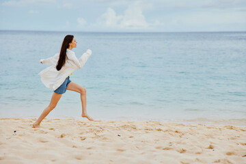 Sports woman runs along the beach in summer clothes on the sand in a yellow T-shirt and denim shorts white shirt flying hair ocean view, beach vacation