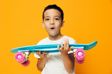 happy, little boy rejoices at the new skateboard in his hands standing on an orange background in a white t-shirt. Horizontal photo with empty space to insert an advertising layout