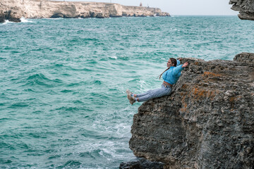 Woman rocks sea. A woman in a blue jacket sits on a rock above a rock above the sea and looks at the raging ocean. Girl traveler rests, thinks, dreams, enjoys nature.