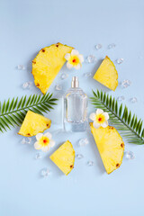 Glass bottle of perfume with pineapple slices, exotic flowers, green palm leaves, ice cubes, water drops on the light blue background. Smell of summer. Fresh pineapple cooling perfumery.