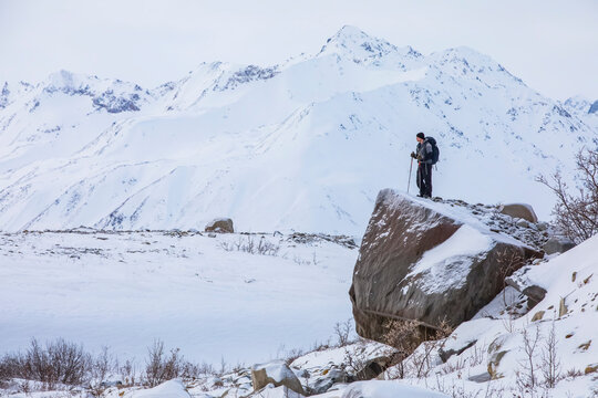 Man hiker stands on an erratic glacial-deposited rock from the retreat of the Black Rapids Glacier overlooking the snowy, winter landscape of the Alaska Range; Alaska, United States of America