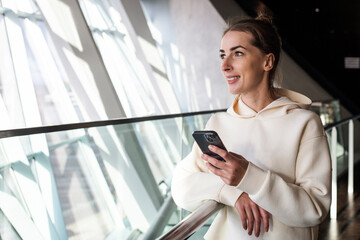 Fototapeta premium Smiling young woman with phone looking at panoramic view from window