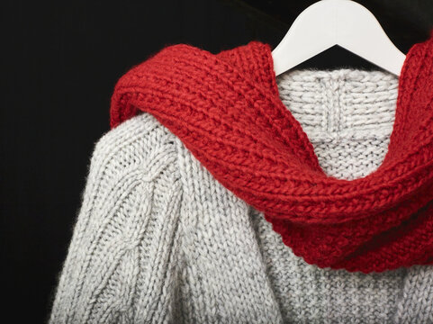 A grey knit sweater draped with a red knit scarf displayed on a hanger on a black background; Studio