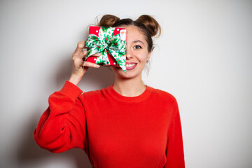 Portrait of young cute brunette girl with two bundles, red sweater smiling widely closing one eye by Christmas red green present, copy space