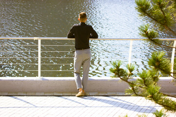 A young handsome slender tall man back view in a black shirt, gray pants stands near a metal fence on a waterfront looking on a river water. Reflection, planning, recreation, business people concept. - Powered by Adobe