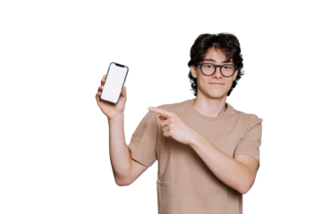 Handsome Italian schoolboy in glasses and beige t-shirt isolated on transparent backdrop pointing at phone with blank screen looks at camera. Cheerful teenager holding smartphone. Mockup. Model.