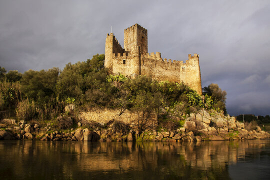 Warm sunlight reflecting on the historical Castlo de Almourol on the islet of Almourol in the River Tagus with a grey sky; Vila Nova da Barquinha, Praia do Ribatejo, Centro Region, Portugal