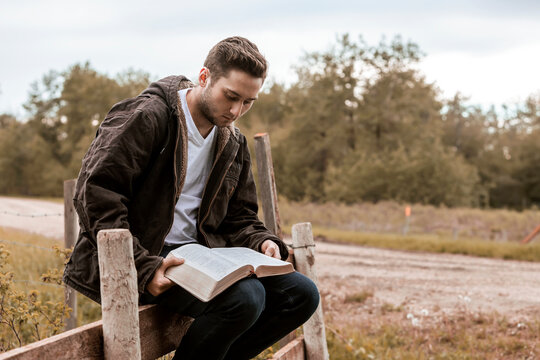 A young man reads from his Bible as he sits on a rail fence in the countryside; Alberta, Canada